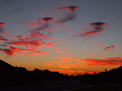 [Above a dark semi-circle of rooftops are horizontal lines of yellow-orange clouds. Above those are pink-purple puffy ones. Several of these have whispy dark masses seeming to come from the bottom as if they are rain which is not coming to the ground.]